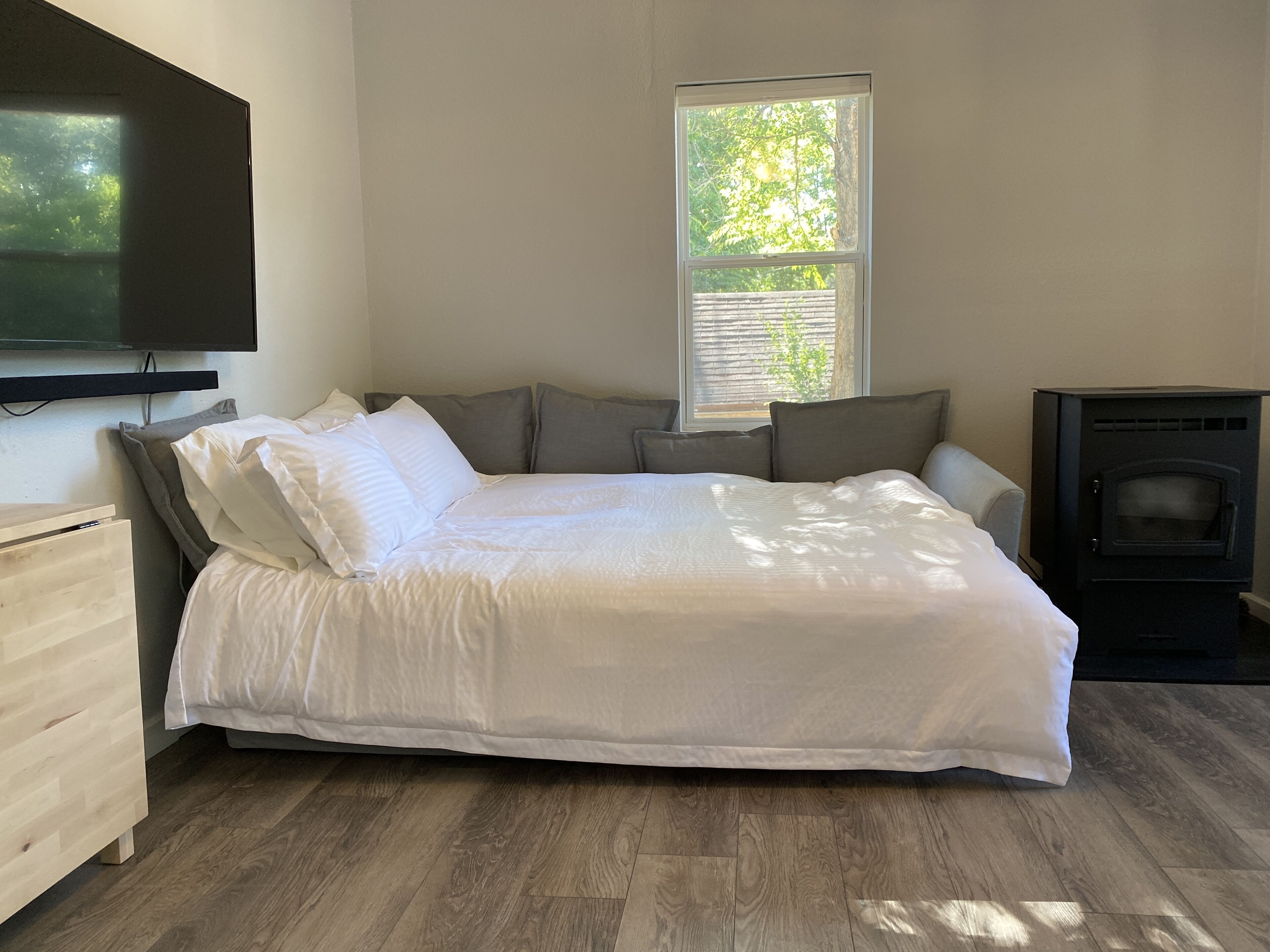 A bedroom with a bed, a TV mounted on the wall, a window with blinds, and a wood-burning stove.