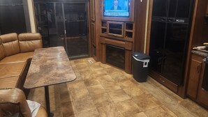 A living room with a brown leather sofa, a small table, a TV mounted on a wooden cabinet, and a trash bin. - Camper on campsite (Jamestown)