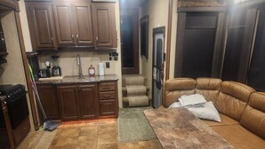 A compact kitchen with wooden cabinets, a countertop, and a refrigerator. There is a brown leather sofa with a pillow and a small table in the living area. - Camper on campsite (Jamestown)
