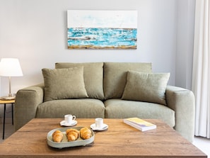 A living room with a green sofa, a wooden coffee table with croissants and cups, and a painting of a seascape on the wall.