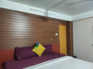 Ein Hotelzimmer mit einem Holzkopfteil, einer violetten Tagesdecke und einem einzelnen gelben Kissen.