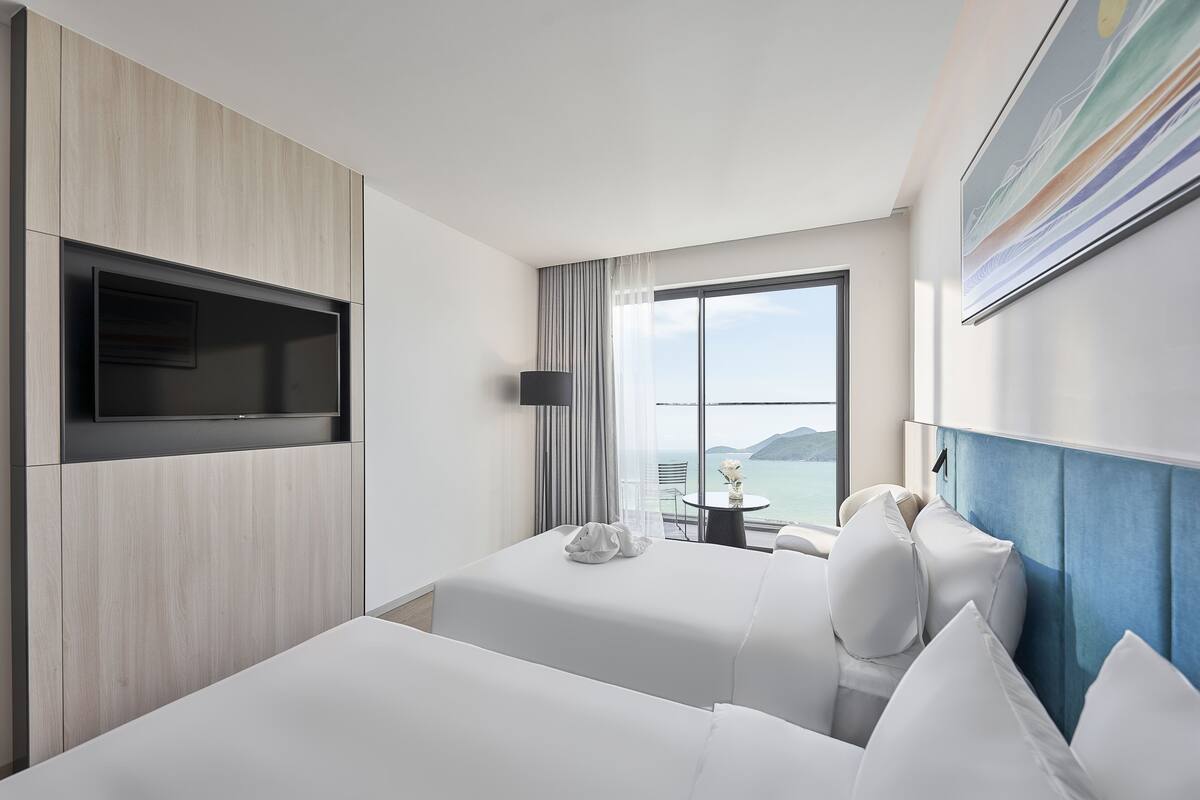 Habitación de hotel con dos camas, un televisor montado en la pared, una mesita con una silla y un ventanal con vistas al mar.