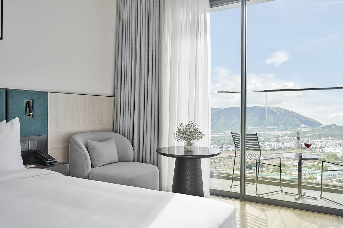 Una habitación de hotel moderna con una cama grande, una mesita redonda con una botella y un vaso, una silla y vistas a una ciudad y montañas a través de ventanas amplias.