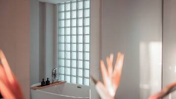 Un baño moderno con bañera, una ventana de diseño entramado y un estante de madera con artículos de aseo.