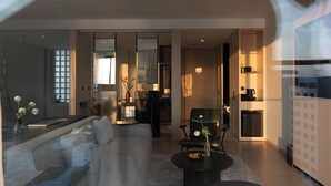A modern living room with a sofa, a coffee table, and a chair. There is a kitchen area with a refrigerator and a coffee machine.