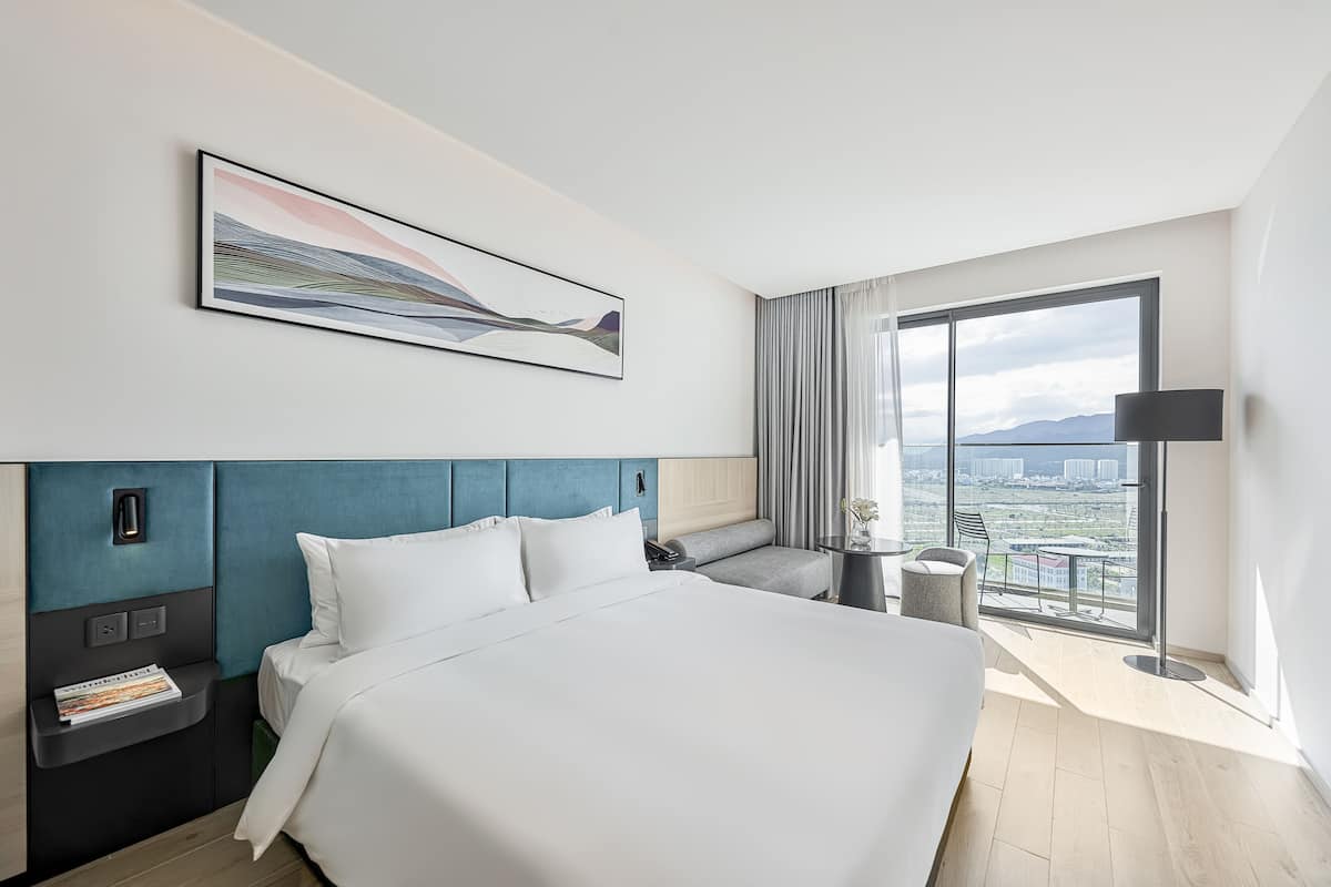 Una habitación de hotel moderna con una cama grande, una mesita de noche, un escritorio, una silla y un ventanal con vistas a la ciudad.