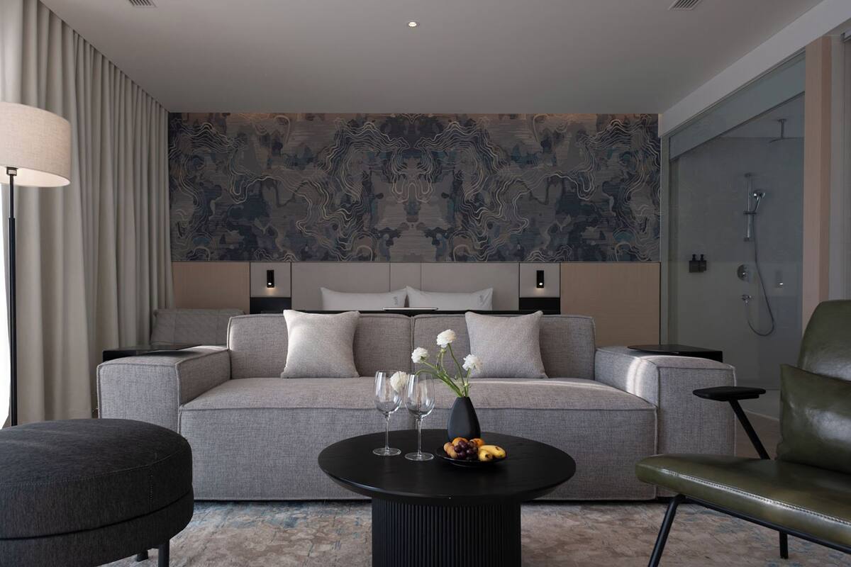 Habitación de hotel moderna con un sofá gris, una mesa de centro negra con un florero y fruta, y una pared grande con diseño.