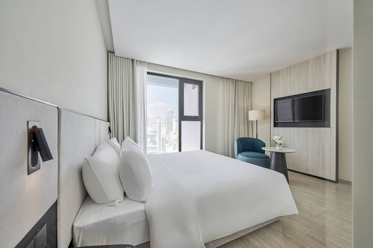 Una habitación de hotel moderna con una cama grande, un televisor montado en la pared, una mesita con una lámpara y vistas a la ciudad desde la ventana.