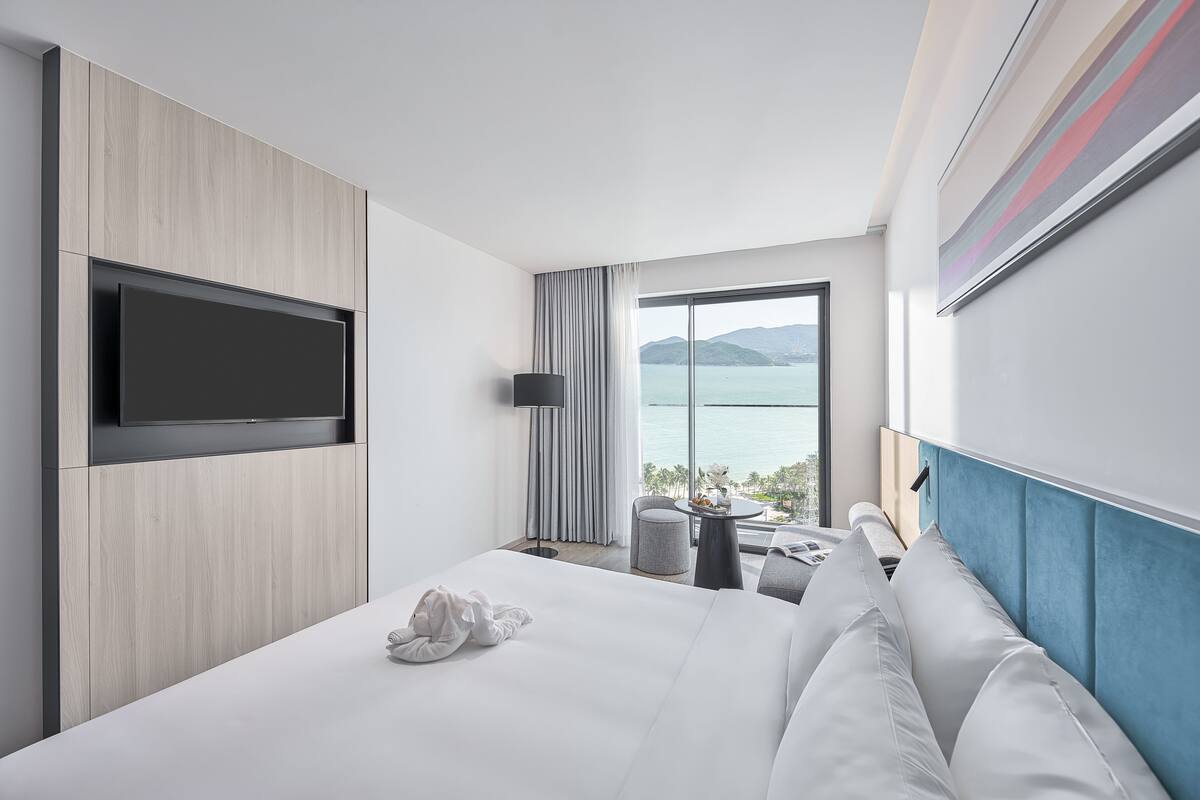 Una habitación de hotel moderna con una cama grande, un televisor de pantalla plana montado en la pared, una pequeña mesa redonda con una lámpara y vistas al mar a través de un amplio ventanal.