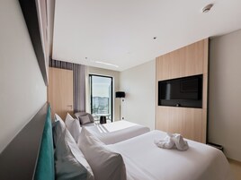 A hotel room with two beds, a TV mounted on the wall, a balcony with a city view, and a small table with a chair.