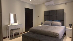 A bedroom with a large bed, a bedside table with a lamp, a dresser, and a door.