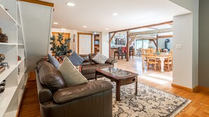 A spacious living room with a brown leather sofa, a glass coffee table, and a patterned rug. There is a dining area with wooden chairs and a table in the background.