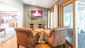A living room with a TV, a round coffee table, two leather armchairs, a wooden cabinet, and a large window with a view of the outdoors.