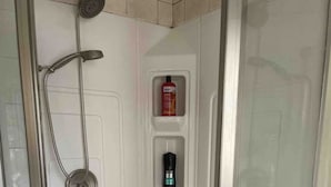 Un box de ducha con paredes de vidrio, equipado con un cabezal de ducha manual y un accesorio de ducha manual, además de un estante empotrado con artículos de tocador.