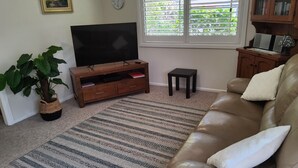 Een woonkamer met een televisie, een bank, een salontafel, een bijzettafeltje, een plant en een raam met jaloezieën.