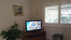 Een woonkamer met een televisie op een houten dressoir, een kamerplant, een klok en een raam met zonwering.