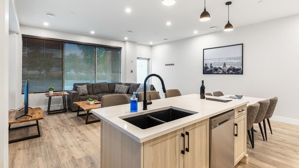 A modern kitchen with a central island, a dining area with a table and chairs, a sofa, and a large window with blinds. - 3-bedroom house Daily/Monthly Rates Available (Parksville)