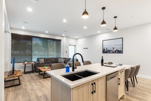 A modern kitchen with a central island, a dining area with a table and chairs, a sofa, and a large window with blinds. - 3-bedroom house Daily/Monthly Rates Available (Parksville)