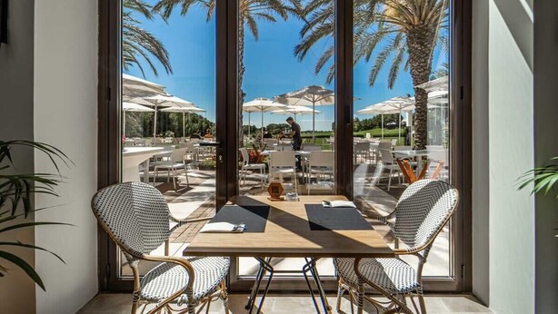 A corner table with wicker chairs, a wooden table, and a view of an outdoor patio with umbrellas and palm trees. - Stunning Penthouse Apartment in Las Colinas - LS18LC (Las Colinas)