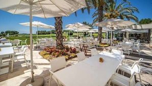 Un espace de restauration en plein air avec des tables et des chaises blanches, entouré de parasols, de palmiers et, au loin, d’un terrain de golf.