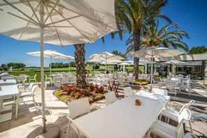 Een terras met witte tafels en stoelen, omgeven door parasols, palmbomen en een golfbaan op de achtergrond.