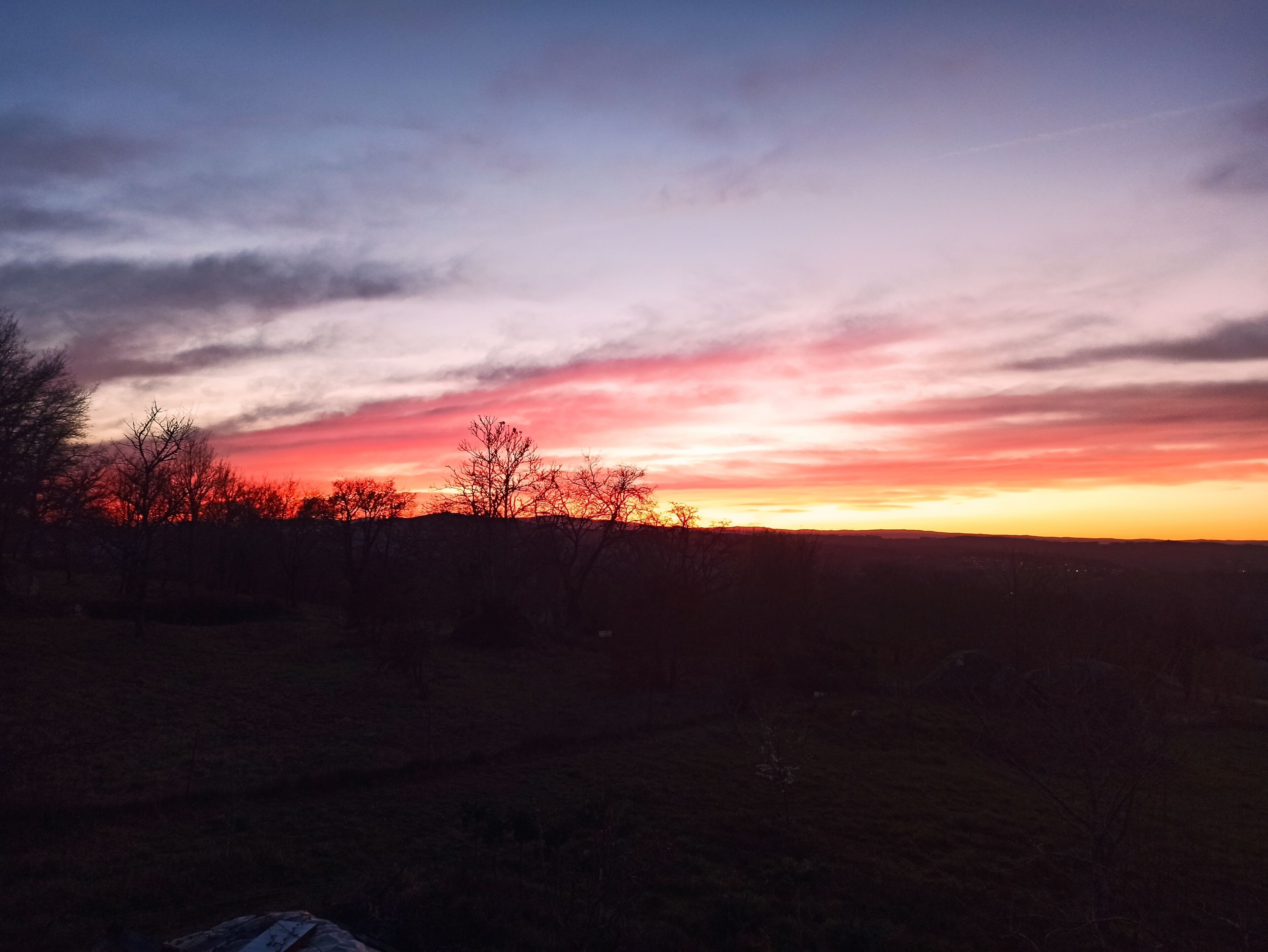 A sunset over a rural landscape with silhouettes of trees and a flat horizon.
