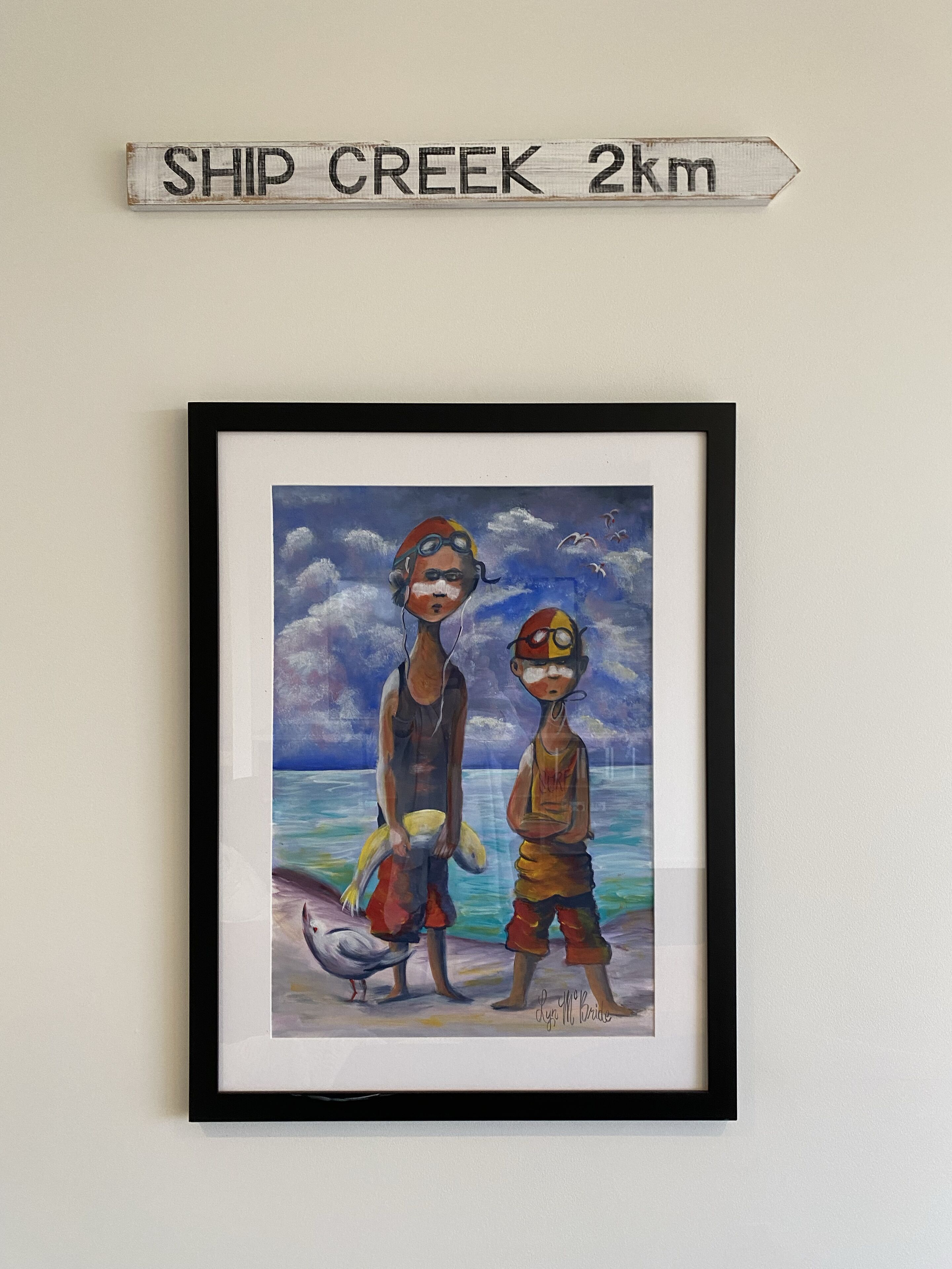 Un tableau encadré représentant deux enfants sur une plage, avec une pancarte au-dessus indiquant « SHIP CREEK, 2 km ».