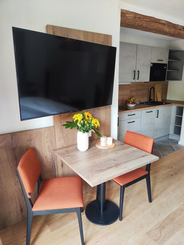 Un espace repas moderne avec une table en bois, deux chaises orange, un téléviseur à écran plat fixé au mur et une cuisine en arrière-plan.