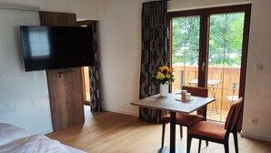A hotel room with a bed, a dining table with chairs, a TV mounted on the wall, and a large window with curtains. - Appartement 1, Benedikt, 28qm (Prien am Chiemsee)