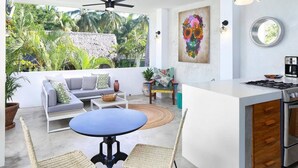 A modern living room with a sofa, a round table, and chairs, a kitchen area with a stove, and a large window with a view of greenery. - Villa Robalo Duplex - Villas del Palmar (Sayulita)