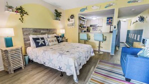 Ein Zimmer mit einem Bett, einem blauen Sessel, einer kleinen Küchenzeile und einem durch eine offene Tür einsehbaren Badezimmer.