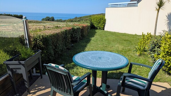 A patio with a round table and chairs, a view of the sea, and a hedge.