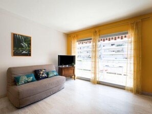 A living room with a sofa, a flat-screen TV, and a large window with yellow curtains.