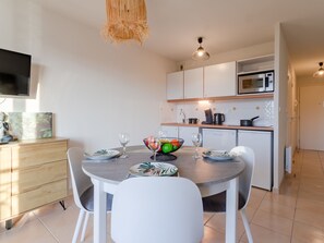 A modern kitchen with white cabinets, a microwave, and a coffee maker. There is a dining table with white chairs and a bowl of fruit.