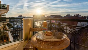 En balkong med ett bord dukat för frukost, med utsikt över en småbåtshamn och stadssiluetten under solnedgången.