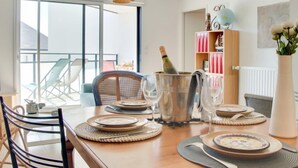 A dining table set with plates, cutlery, and a champagne bottle, with a wicker chair and a hanging light fixture in the background. - Stella Marine by Interhome (Carnac)