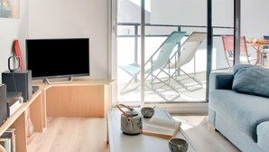 A modern living room with a flat-screen TV, a sofa, a coffee table with a book and cups, and a balcony with chairs. - Stella Marine by Interhome (Carnac)