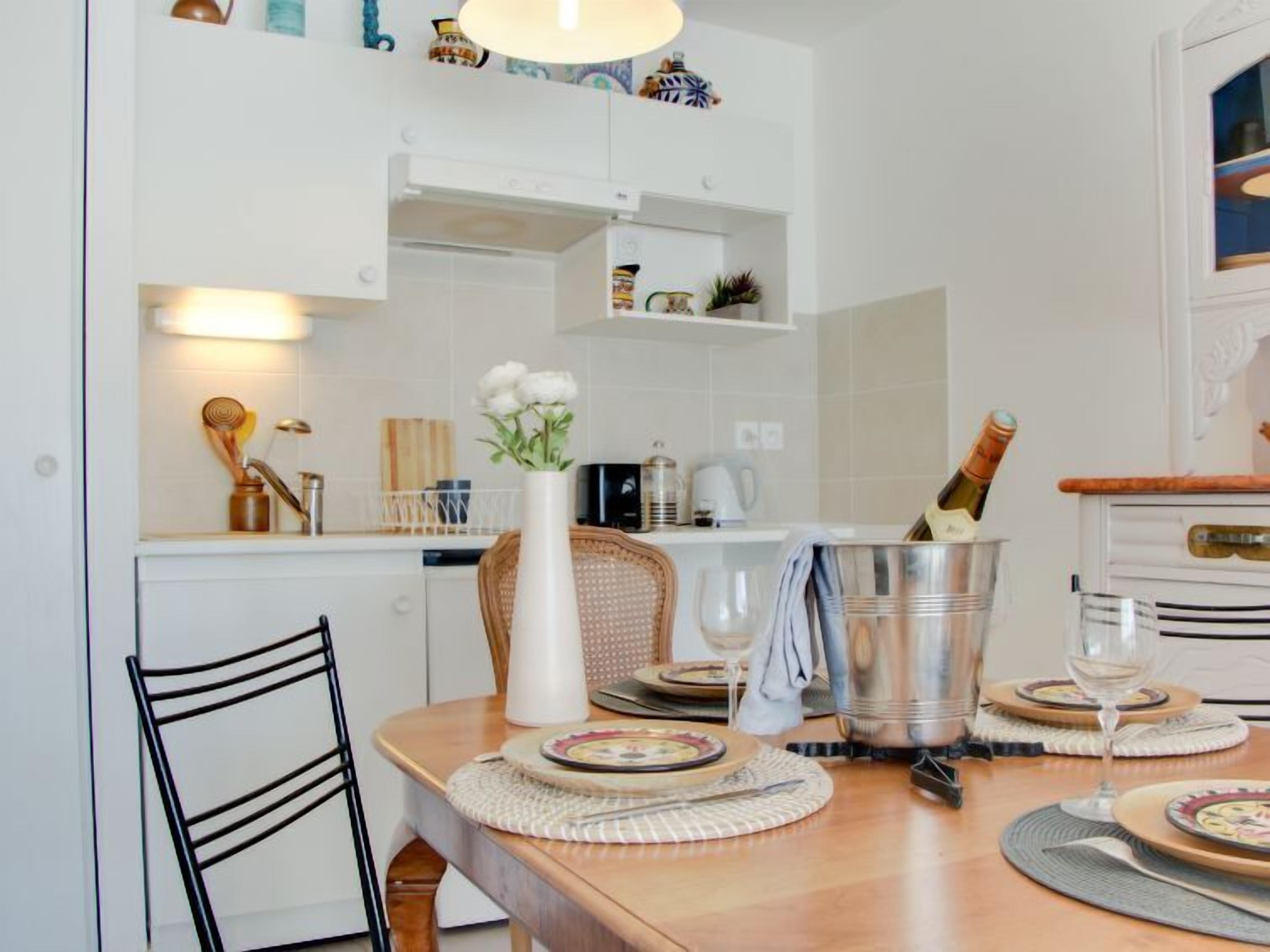 A dining area with a wooden table set for two, a vase with flowers, a bottle of champagne, and a buffet bar in the background.