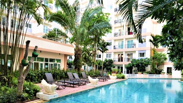 A swimming pool with lounge chairs, surrounded by palm trees and a multi-story building with balconies. - 1 bed Condo With Direct Pool Access - Sleeps 2 (Pattaya)