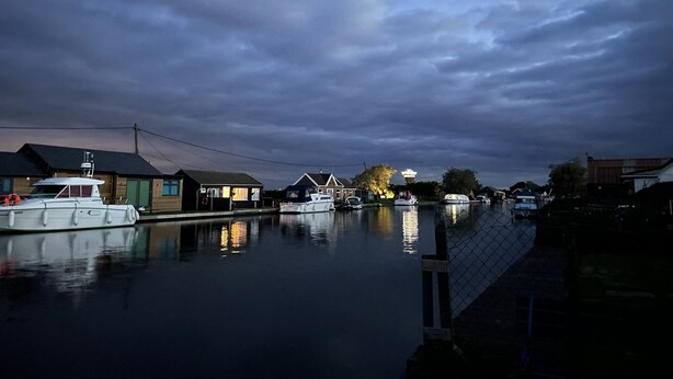 A calm river with boats docked on the left, surrounded by houses and a cloudy sky.