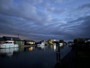 A calm river with boats docked on the left, surrounded by houses and a cloudy sky.