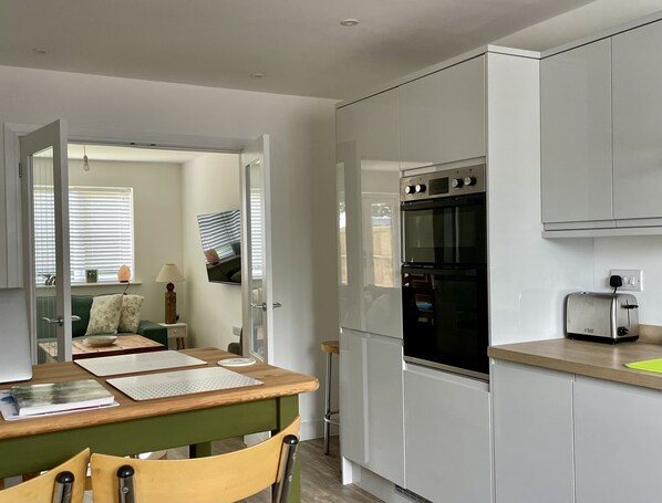 A compact kitchen with white cabinets, a double oven, and a toaster. There is a dining table with chairs and a window with blinds.
