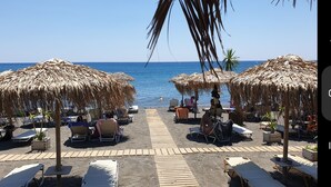 Ein Strandabschnitt mit Reetdachschirmen, Liegestühlen und einem Holzsteg, der zum Meer führt.