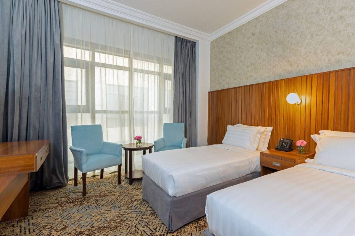 A hotel room with two beds, a desk, a chair, a small table, and a nightstand with a lamp.