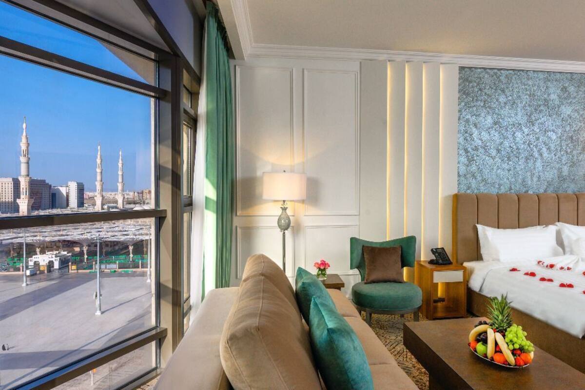 A hotel room with a large bed, a sofa, a chair, a small table with fruit, and a view of a cityscape with buildings and a bridge.