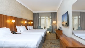 Ett hotellrum med två sängar, ett skrivbord, en stol, en lampa och ett stort fönster med gardiner.