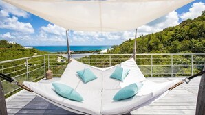 A hammock with cushions on a balcony overlooking a scenic view of a coastline and greenery.