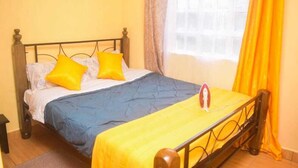 A bed with a blue and yellow bedspread, two yellow pillows, and a red iron on the bed. - Stay.Plus Makey Cozy Homes (Ngong)
