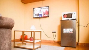 A living room with a television mounted on the wall, a small wooden table, a refrigerator, and a sofa. - Stay.Plus Makey Cozy Homes (Ngong)