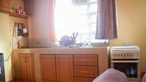 A compact kitchen with wooden cabinets, a stove, and a window with curtains. - Stay.Plus Makey Cozy Homes (Ngong)